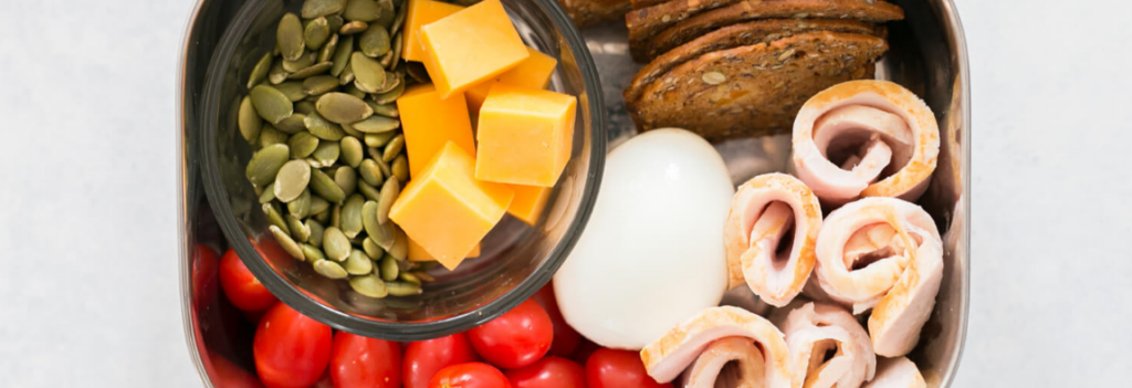 A bento box of hard-boiled egg, crackers, cherry tomatoes, seeds, cheese and sliced turkey.